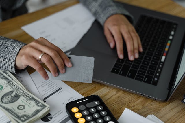 Person using a laptop while holding a bank card, money and a calculator are sitting on the table next to the laptop